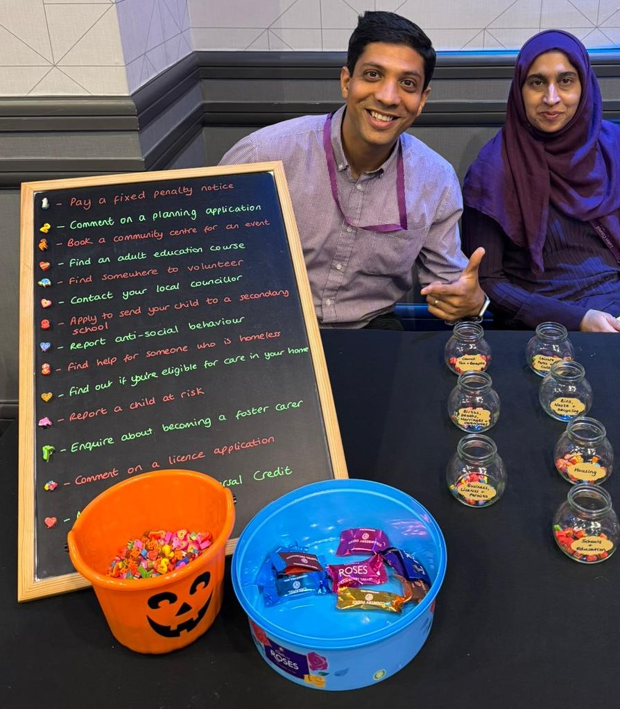 Two people sitting down next to each other behind a table. The table has a black cloth and a chalkboard with jars on top of the table.